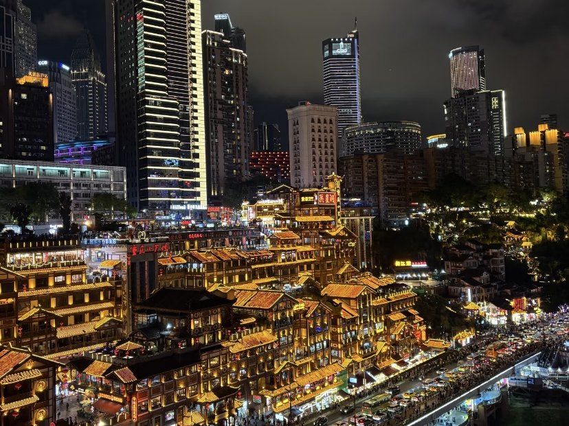 Hongya Cave lit up gold at night, with high-rises behind and traffic on the riverside road